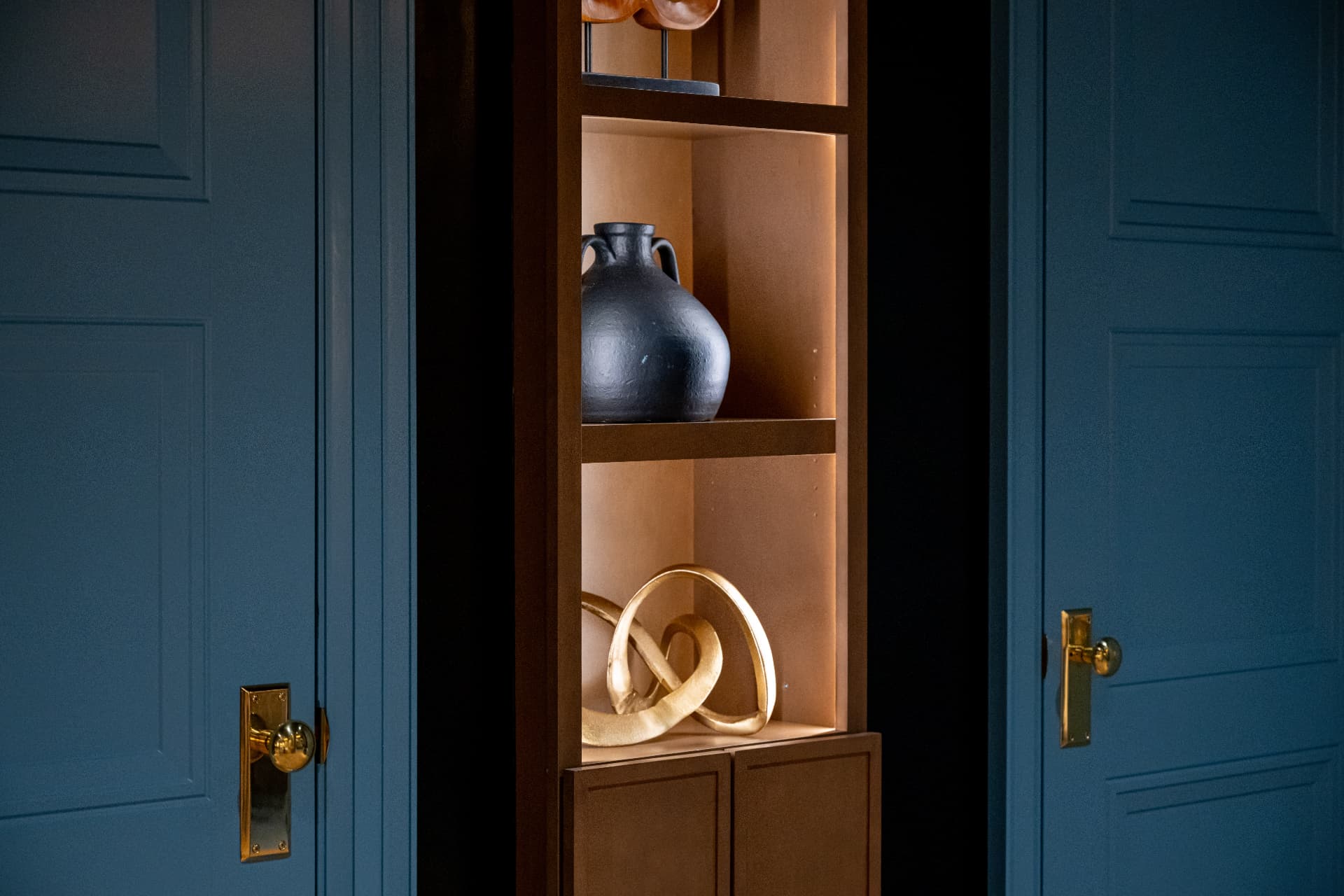 Illuminated wood shelving with ceramic vessels between deep blue doors and brass hardware
