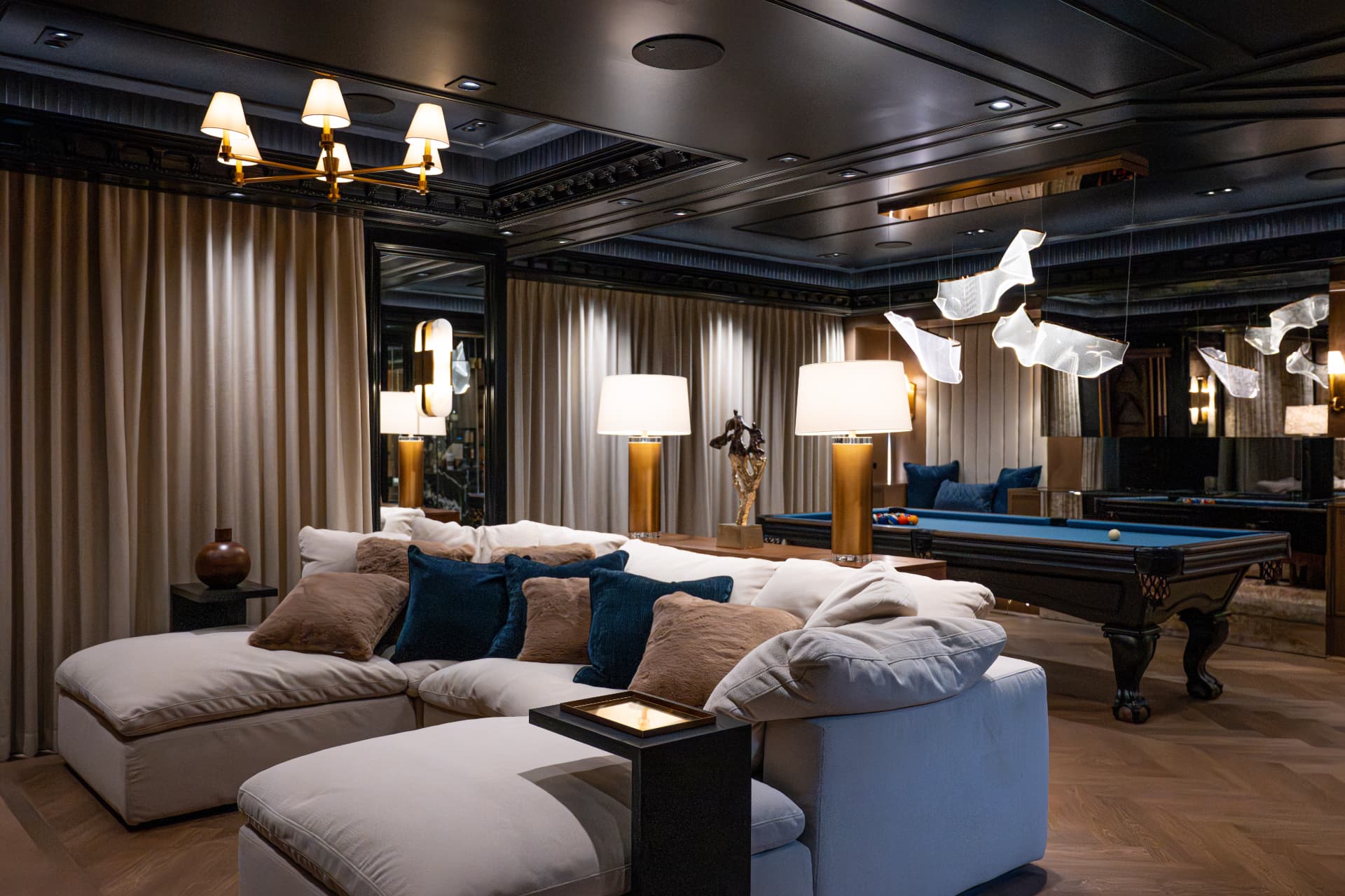 Open lounge with a cream sectional, layered pillows, brass lamps, and a pool table beneath sculptural chandeliers