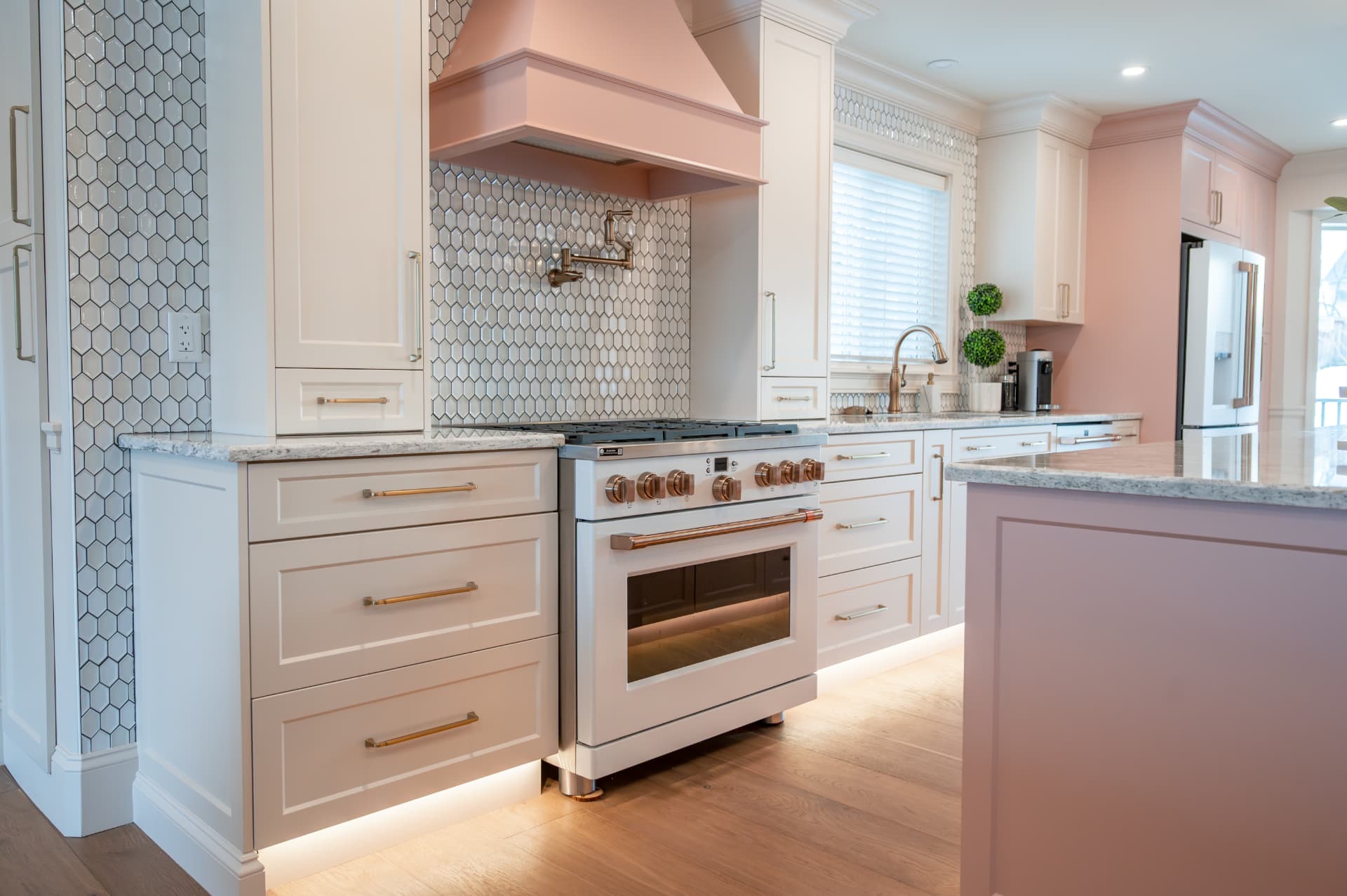 Range with copper hardware set against a hexagon tile backsplash and softly lit blush cabinetry