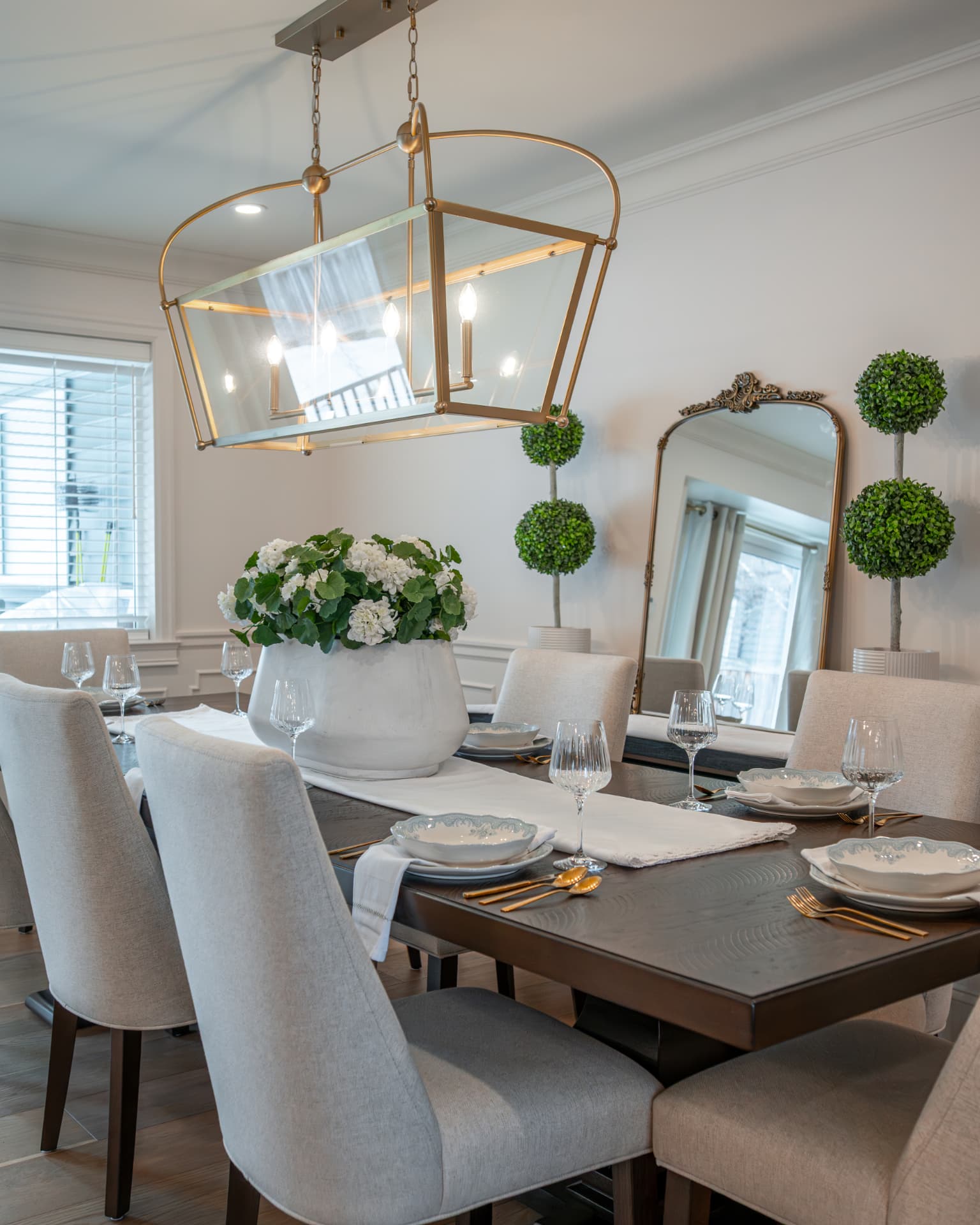 Dining table set for entertaining beneath a brass and glass linear chandelier with white hydrangeas