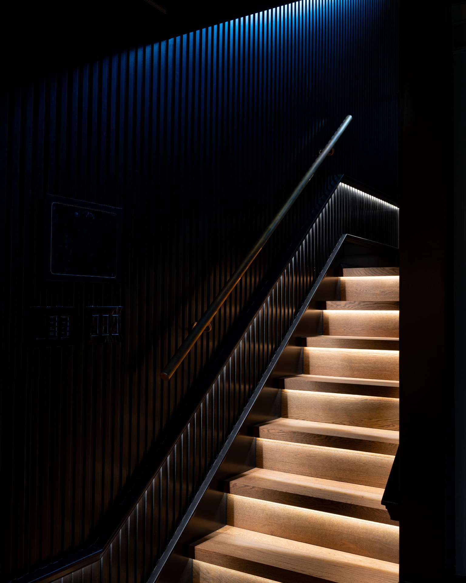 Dark stairwell with illuminated wood treads, black fluted walls, and a brass handrail
