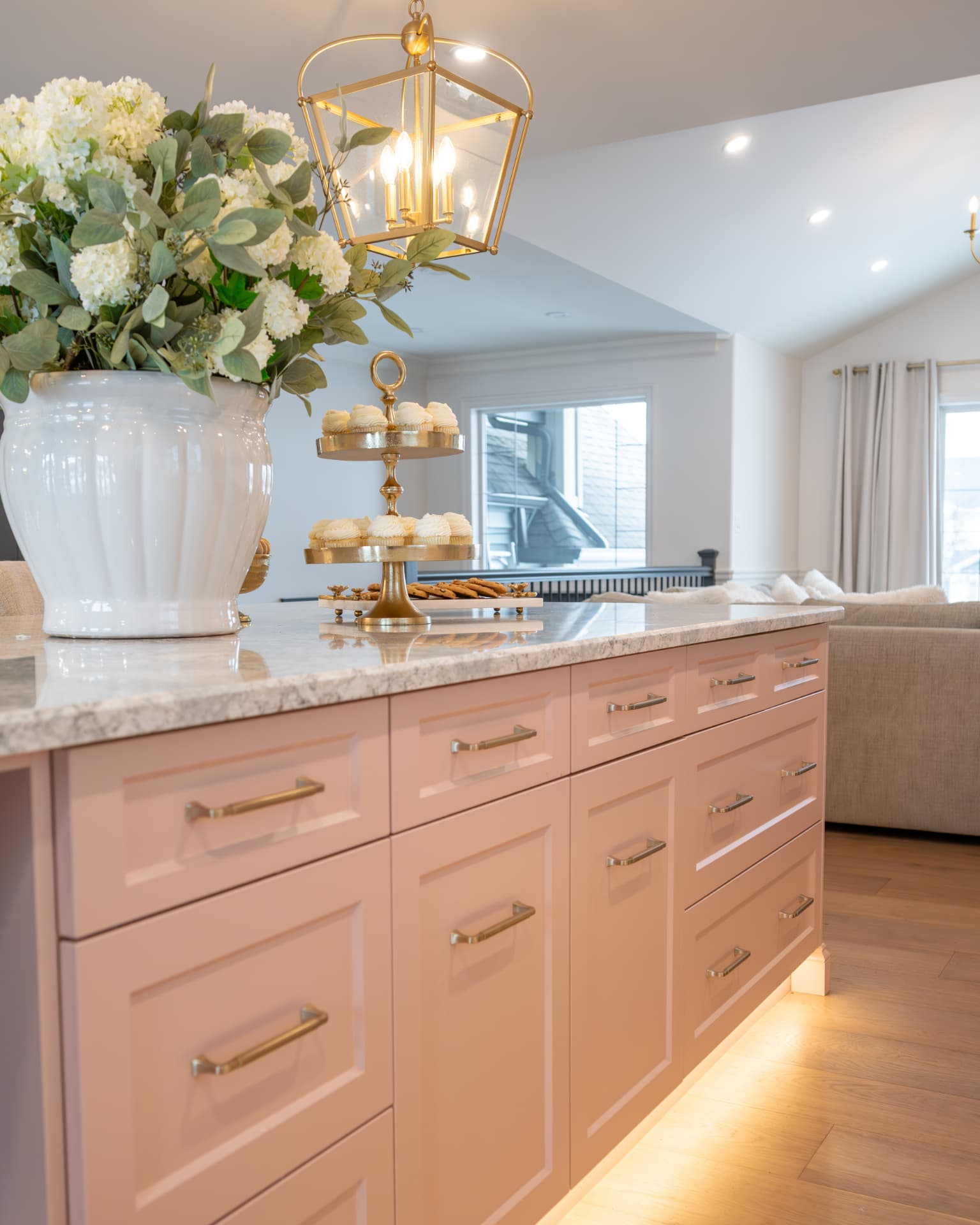 Blush island counter with a marble surface, gold dessert stand, and white florals beneath a brass lantern