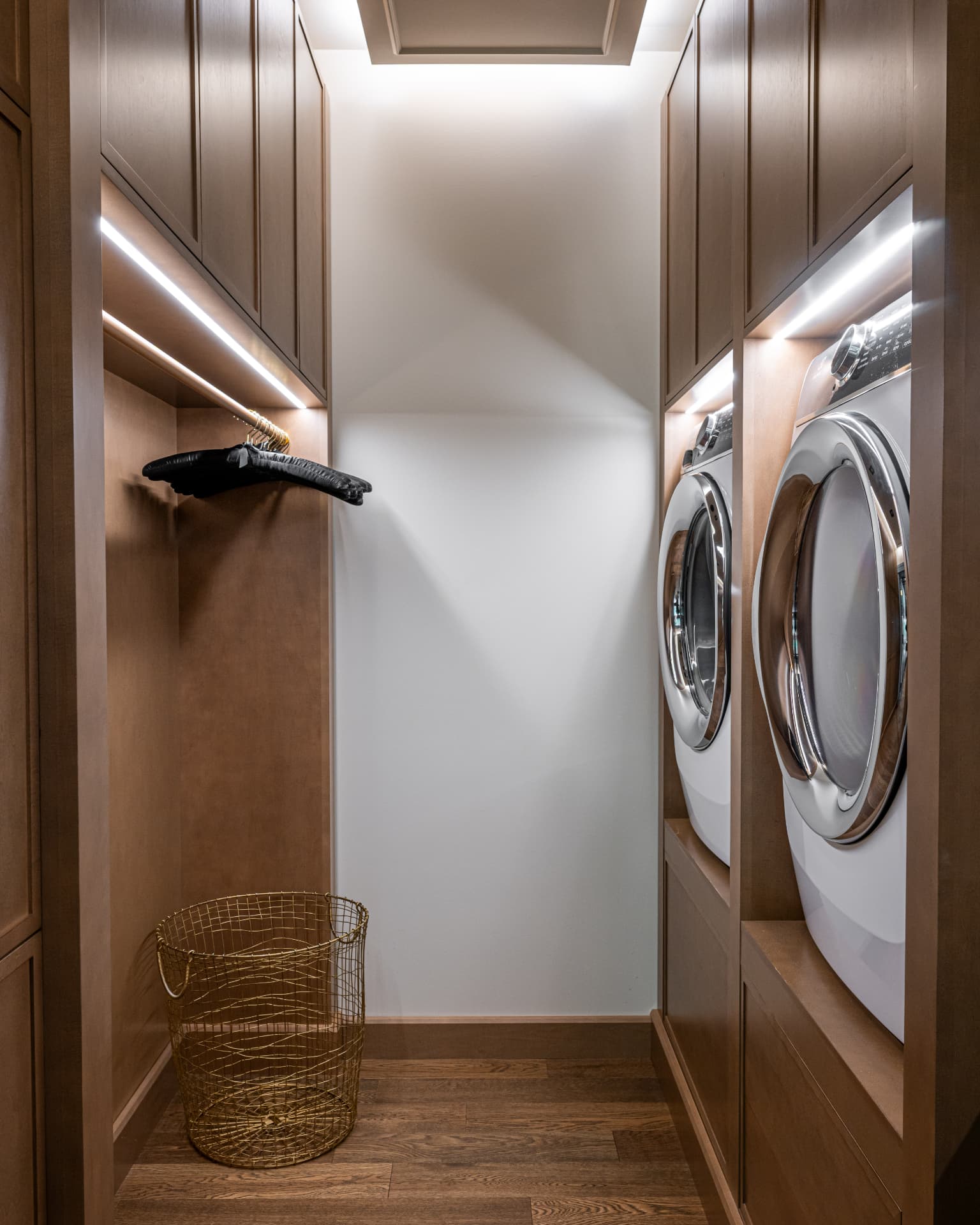 Tall custom laundry cabinetry with integrated washer and dryer, illuminated hanging rail, and brass basket