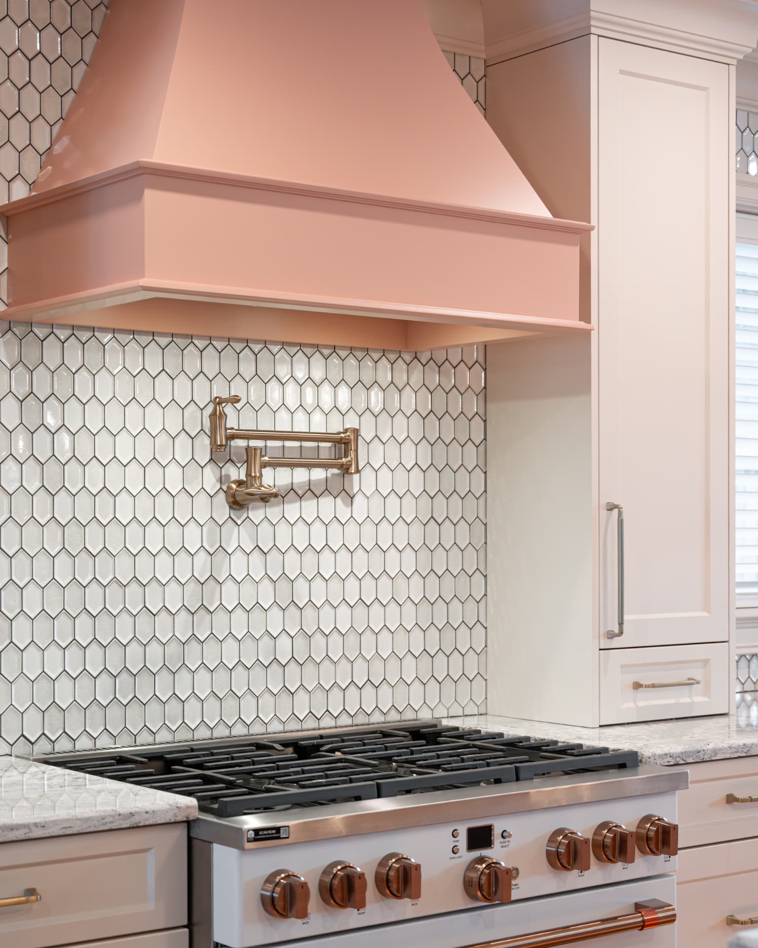 Copper range hood above a marble hexagon backsplash with a brass pot filler and illuminated under-cabinet lighting