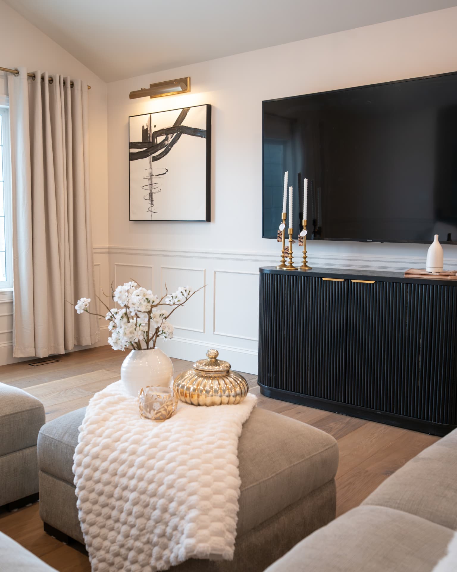 Family room with a black fluted media cabinet, brass picture light, framed art, and cream furnishings