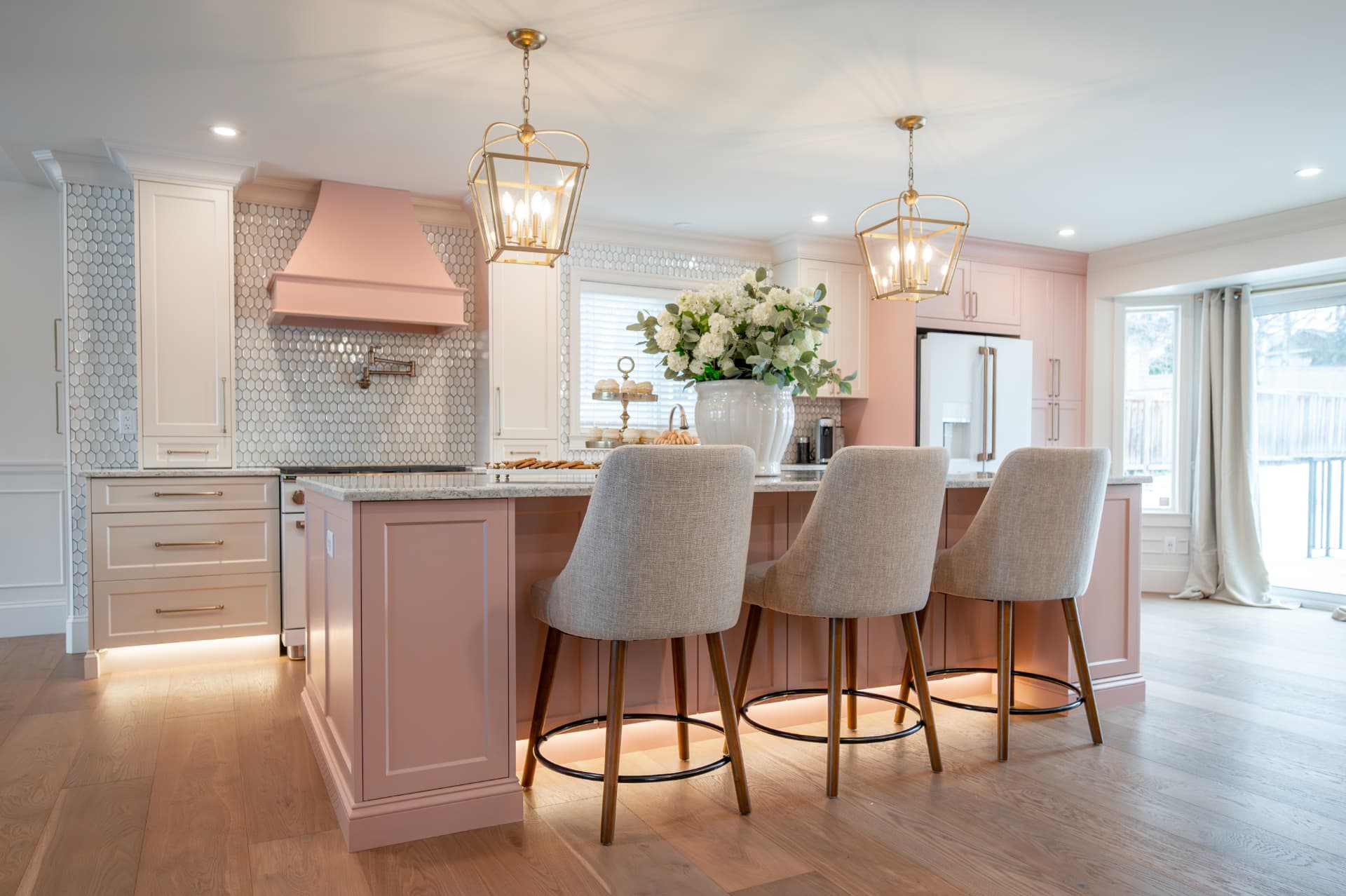Kitchen island in blush cabinetry with three upholstered bar stools, brass lantern pendants, and fresh hydrangeas