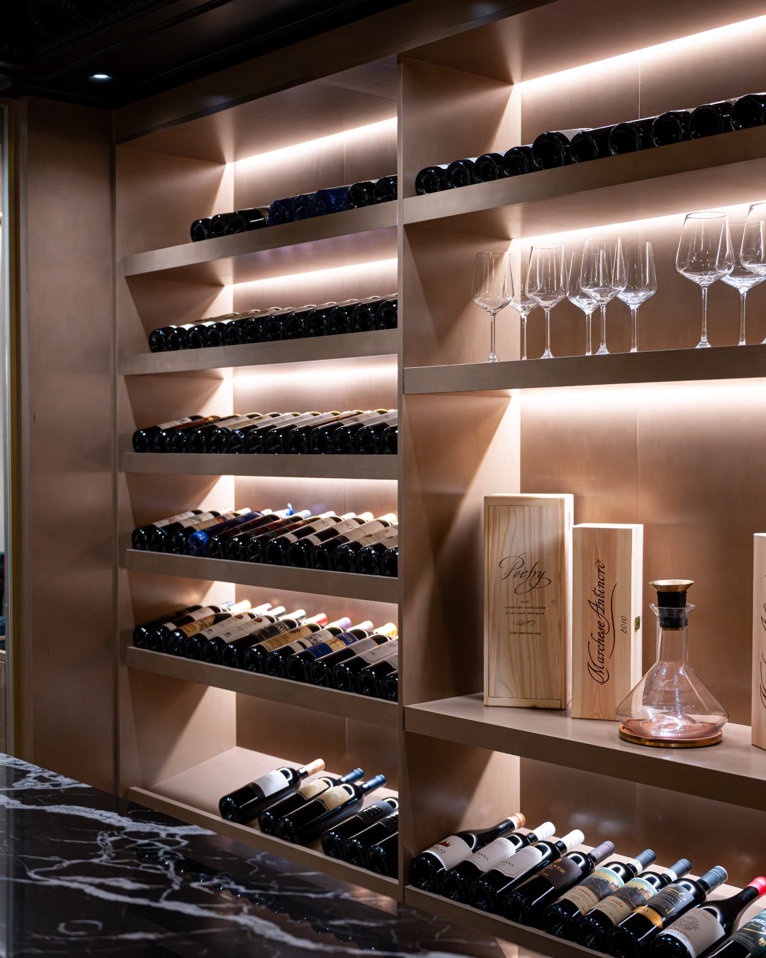 Illuminated wine cellar shelving lined with bottles, stemware, and wooden cases