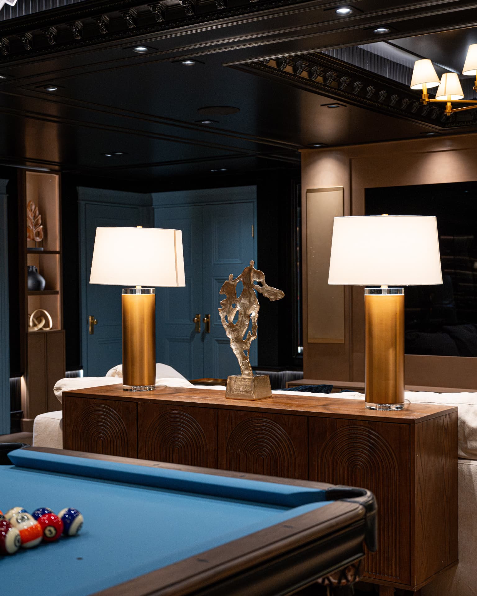 Brass column lamps and a sculptural figure atop a carved wood console beside a blue pool table